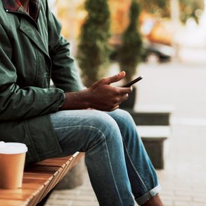 Person sitting with smartphone on a bench, outdoor setting. Nearby, a paper cup, camera, and backpack on the bench.
