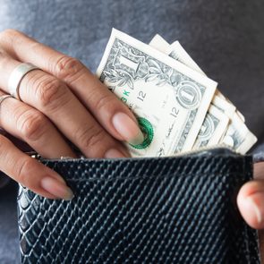Hands hold a black textured wallet, inserting dollar bills, with a person in a gray sweater nearby.
