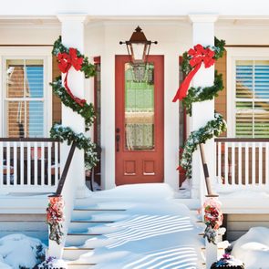 Red door with glass panel, adorned with Christmas garlands and bows, snow-covered steps, white railing, brown house.