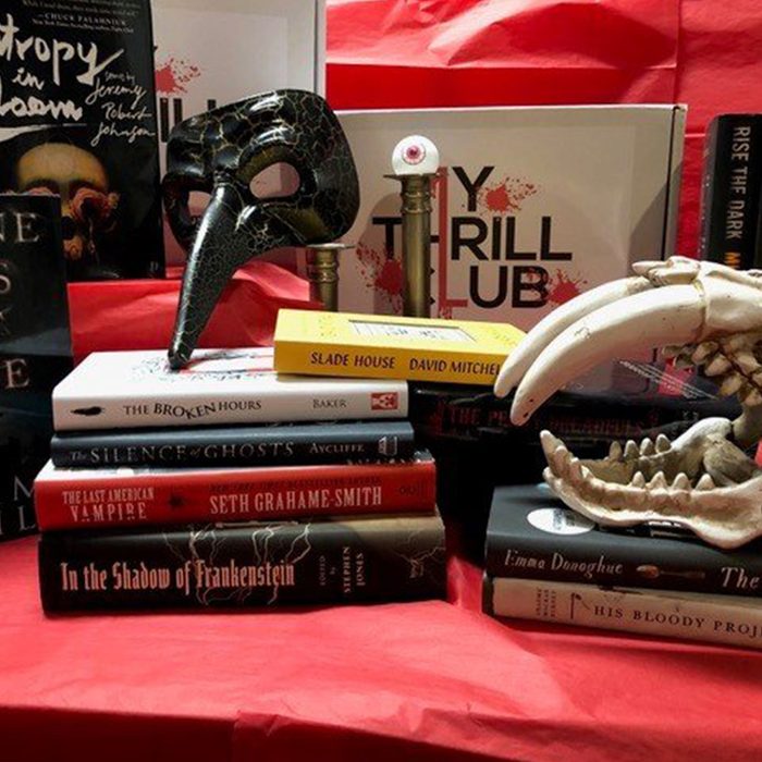 A stack of mystery novels topped by a plague mask and a candle, set against a red backdrop with a skull decoration.