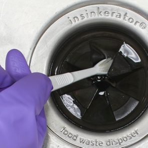 Gloved hand scrubs sink's food waste disposer opening with a toothbrush, cleaning black rubber flaps. 