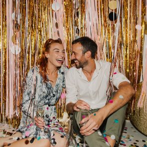 A couple laughs, holding wine glasses, surrounded by sparkling gold and pink streamers and scattered confetti.