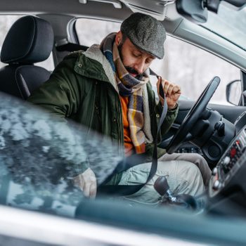 Man wearing a flat cap and scarf fastens seatbelt inside a parked car, surrounded by snowy landscape visible through the windows.