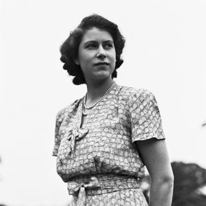 A woman stands looking sideways, wearing a patterned dress and pearl necklace, outdoors under a bright, clear sky.
