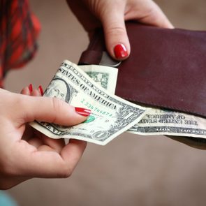 Hands hold a leather wallet, pulling out U.S. dollar bills; background features blurred plaid clothing.