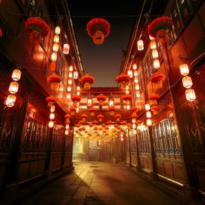 Lanterns illuminate, hanging in rows, creating a warm glow between traditional wooden buildings in an alleyway during nighttime.