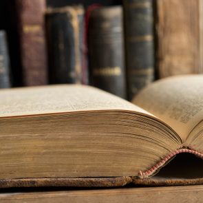 An open old book rests on a wooden surface, with several more aged, leather-bound books standing upright in the blurry background.
