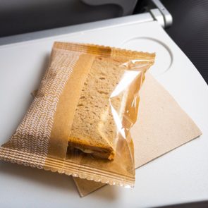 A packaged sandwich rests unopened on an airplane tray table with a napkin beneath it.