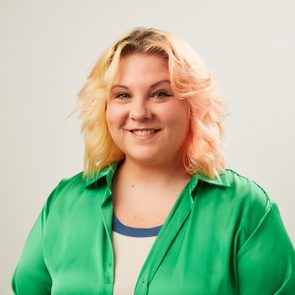 A smiling person poses against a plain backdrop, wearing a green shirt over a beige top, with light, wavy hair.
