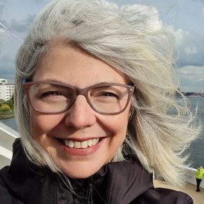 A smiling person with gray hair wearing glasses, stands outdoors, with water and buildings in the background, under a partly cloudy sky.