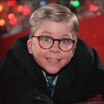 Child smiling, wearing glasses, lying on a red slide. Background features blurred colorful Christmas lights.