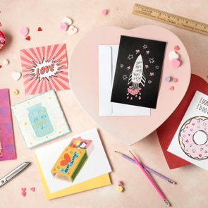 Greeting cards are arranged on a pink surface with heart-shaped candies and pencils; messages express themes of love and friendship.