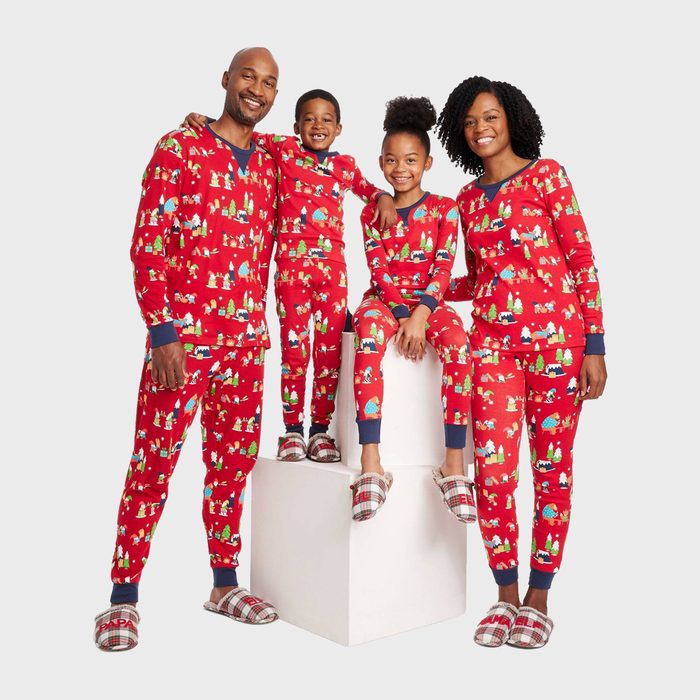 Four people wear matching red holiday pajamas, standing and sitting on white cubes, smiling warmly in a studio setting with plain background.