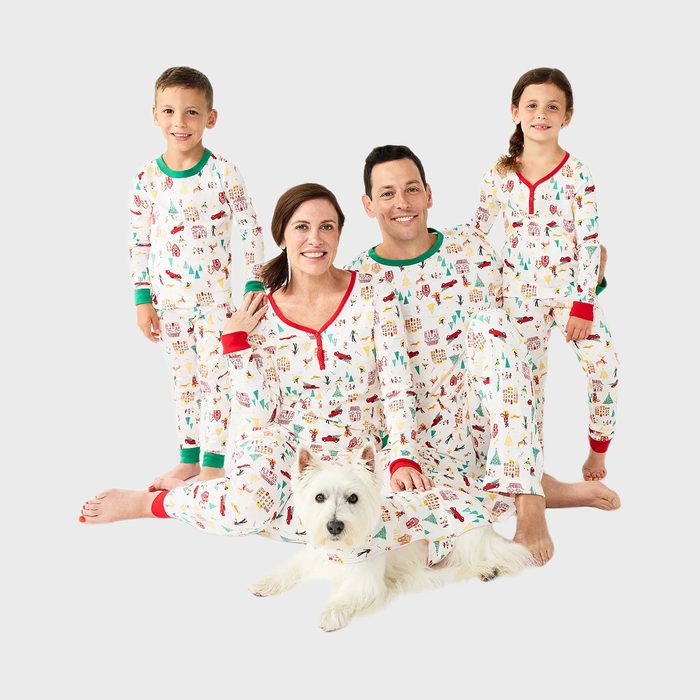 Family wearing matching festive pajamas, sitting together on the floor, smiling. A small dog in similar pajamas is seated in front.