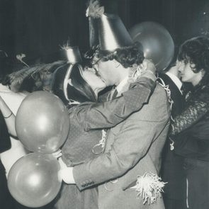 A couple kisses, wearing party hats and holding balloons, surrounded by others at a festive gathering.