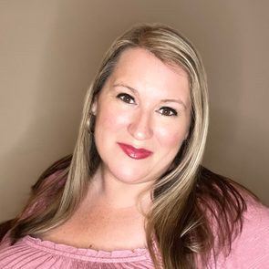 A person smiles softly, wearing a pink top, framed by light brown hair, against a plain beige background.