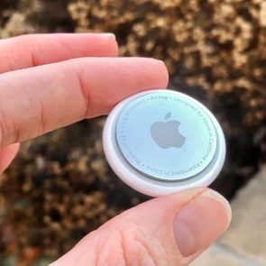 A hand holds a round, white device with an Apple logo. The background is blurred, earthy tones. Text reads: â€œAirTag - Designed by Apple.â€