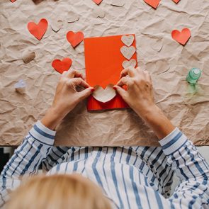 Hands glue hearts on a red card, surrounded by paper hearts on crumpled brown paper. A person wears a striped shirt.