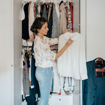 Woman holds a white blouse, standing beside an open wardrobe filled with various clothes in a bright room.
