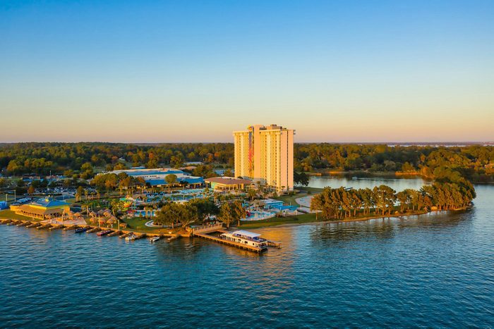 Tall building stands surrounded by water, trees, and boats in a scenic lakeside resort setting under a clear blue sky.