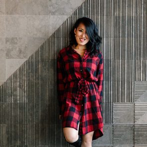 Person in a red plaid dress leans casually against a textured, geometric-patterned wall, smiling with hair slightly tousled.