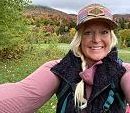 A person smiles, taking a selfie in a field with green and autumn-colored trees under a cloudy sky.