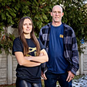 Two people stand confidently outside, surrounded by greenery and a concrete fence, wearing casual shirts.