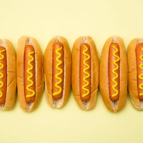 Six hot dogs with mustard line up neatly on a pale yellow background.