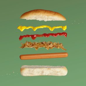Hot dog components float, separated: bun, mustard, ketchup, onions, sausage. Set against a plain green background.