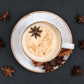 A cup of chai latte topped with star anise and cinnamon, resting on a saucer with spices scattered on a dark surface.