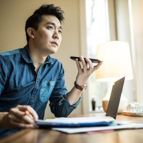 Person holding a smartphone, speaking into it; seated at a desk with notebooks, laptop, and lamp in a well-lit room.