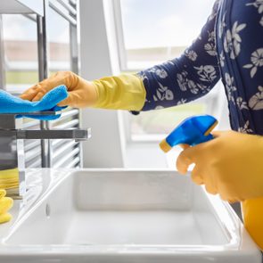 Person cleans a bathroom faucet using a blue cloth and spray bottle, wearing yellow gloves beside a white sink.