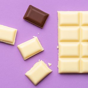 White chocolate bar partially unwrapped with four pieces broken off, one dark chocolate, on a purple surface.