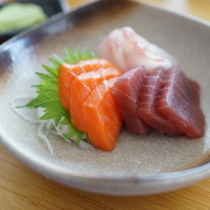 Sashimi slices rest on a ceramic dish with a garnish leaf, on a wooden table, accompanied by a small black dish of wasabi.