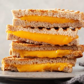 Grilled cheese sandwiches stacked on a plate, with melted cheese oozing between toasted bread slices, gray cloth nearby.