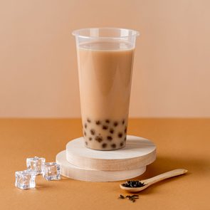 A plastic cup of bubble tea rests on wooden coasters, surrounded by ice cubes and a wooden spoon with tea leaves on a brown surface.