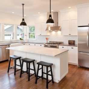 Kitchen with white cabinets and stainless steel appliances, featuring a central island with stools. Sunlight illuminates through large windows, highlighting wooden floors.
