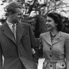 A man and woman walk together, smiling at each other in a garden with trees in the background. Both wear formal suits.