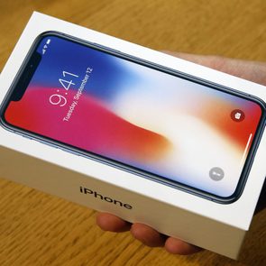 A hand holds an iPhone box displaying the phone's screen at 9:41 AM, against a blurred wooden surface background.