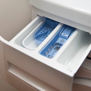 Washing machine detergent drawer open, revealing three compartments with blue inserts, in a bright, neutral-colored laundry area.