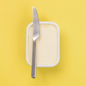 A butter knife rests on an open tub of smooth margarine, set against a solid yellow background.