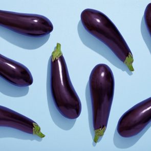 Eggplants are displayed scattered against a light blue background, showing vibrant purple skin and green stems.