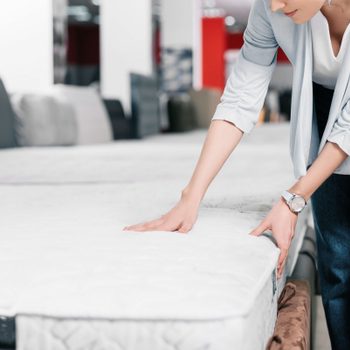 A person presses a mattress with their hands, in a store displaying various bedding products in the background.