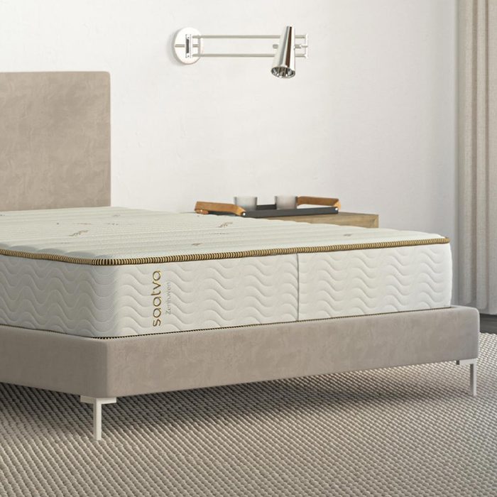 Mattress rests on a beige bed in a modern bedroom; wall lamp and tray on nightstand nearby. Text reads 