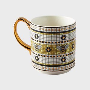 A mug features bee and floral designs on a mosaic-like pattern, with a shiny gold handle, against a plain background.