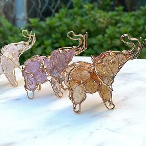 Wire-formed elephants stand on a marble surface, displaying translucent gem bodies, set against a backdrop of lush green foliage.