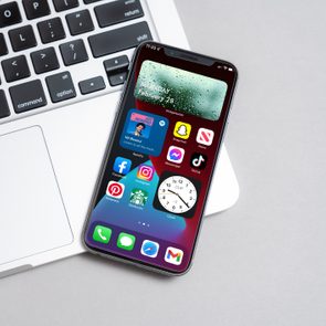 Smartphone displaying app icons and clock lies on a laptop keyboard in an office setting. The screen shows 