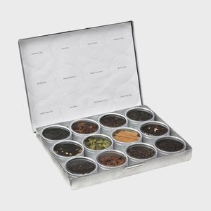 Metallic box holding twelve spice tins, each with visible herbs or spices. Lid displays labeled circles like 