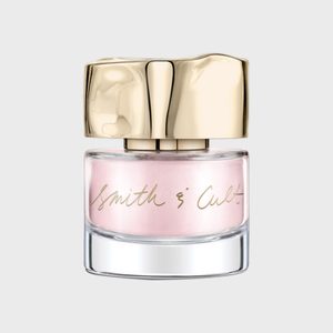 A glass nail polish bottle displays a light pink hue, featuring a gold, reflective cap. The label reads 