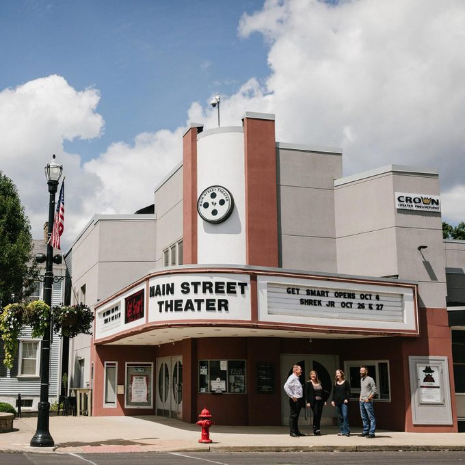 Theater displays show listings on its marquee, beneath a clear sky, with people gathered at the entrance and a nearby streetlamp with hanging plants.